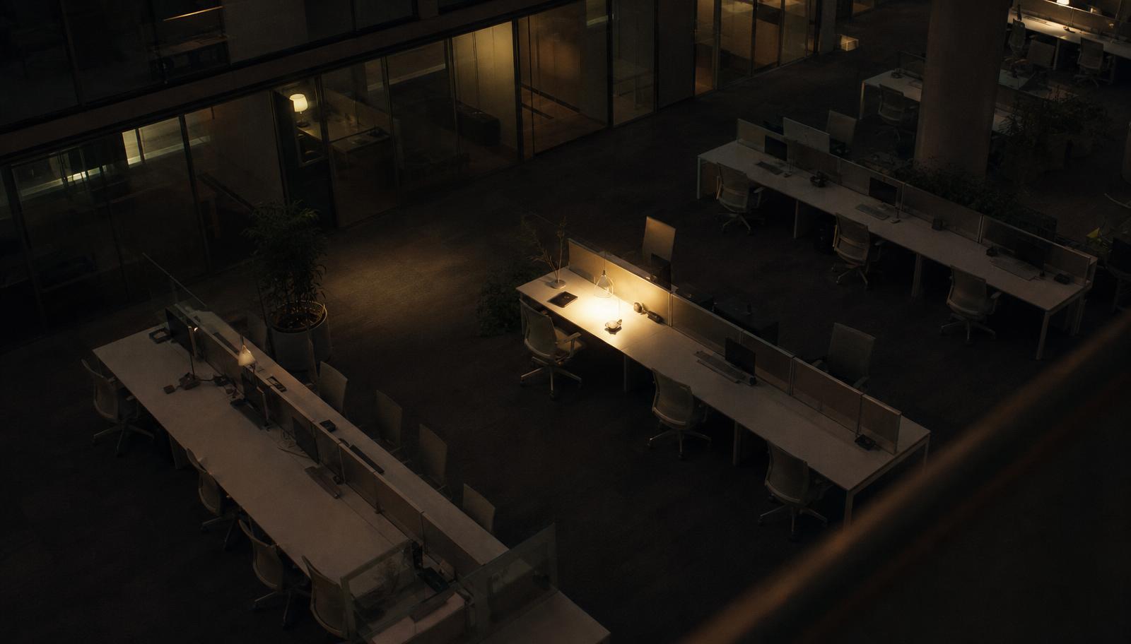 An open-plan office at night seen from above, nearly every desk empty and one lamp still lit